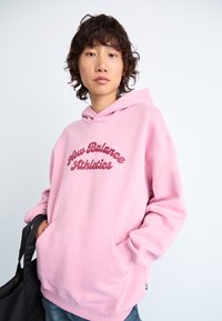 Personne portant un sweat à capuche New Balance Athletics rose, les mains dans la poche kangourou, tenant une sangle de sac noire, face à l'avant sur un fond blanc.