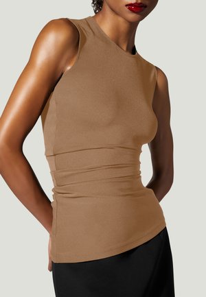 Kvinde iført ærmeløs, tætsiddende brun top og sort underdel, med rød læbestift og kort krøllet hår, foran en bleggrå baggrund.