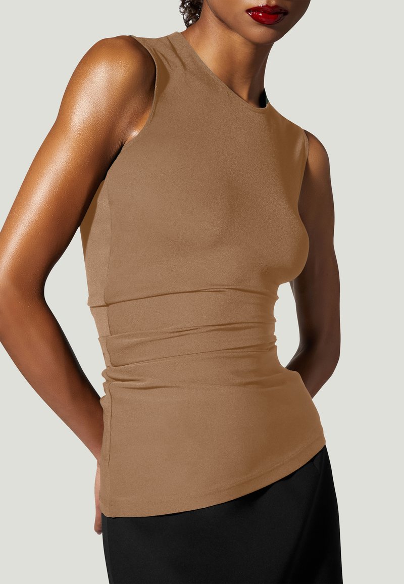 Frau trägt ärmelloses, tailliertes, beige Oberteil und schwarze Hose, mit rotem Lippenstift und kurzen lockigen Haaren, vor hellgrauem Hintergrund.