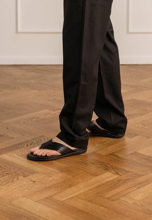 Persoon draagt zwarte broek en zwarte leren sandalen, staand op een lichte houten parketvloer binnenshuis.