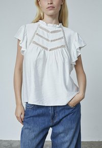 Hvid kortærmet bluse med flæsekant, der har et struktureret chevronmønster og en sammensamlet plissédesign. Matchende med blå denimjeans.