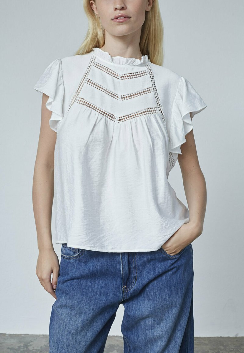 Hvid kortærmet bluse med flæsekant, der har et struktureret chevronmønster og en sammensamlet plissédesign. Matchende med blå denimjeans.