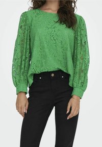 Femme portant un chemisier en dentelle vert vif à manches longues bouffantes et un jean noir taille haute, debout devant un fond uni.