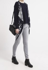 Svart och vit mönstrad cardigan med fransdetaljer, bärs över en mörk tröja och ljusgrå skinny jeans. Svarta ankelboots och en svart väska.