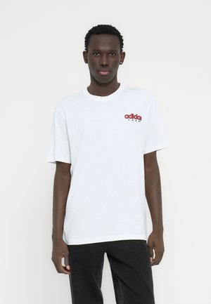 Wit katoenen T-shirt met een rood 'adidas' logo op de borst, korte mouwen en een ronde halslijn. Draagt donkere spijkerbroek.