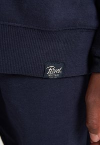 Sudadera de color azul marino con puños de canalé. Una etiqueta negra dice "Petrol Industries" en dorado, cosida en el lado izquierdo cerca del dobladillo.