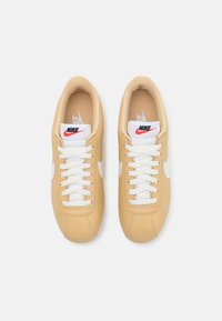 Paire de baskets Nike beige avec lacets blancs et logos swoosh blancs, vue de dessus sur un fond clair uni.