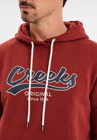 Sudadera roja hecha de una mezcla de algodón, con un logo "Creeks" en azul marino y cordones blancos con extremos de metal. Detalle de escote acanalado.