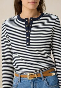 Marineblauw en witte gestreepte langetermijn T-shirt met een ronde hals en vijf gouden knoopsluiten aan de sluiting. Gemaakt met een tankleurige riem.
