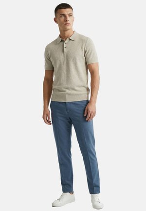 Mężczyzna stojący, ubrany w beżową koszulkę polo z krótkim rękawem, niebieskie spodnie wąskie i białe tenisówki na jednolitym tle.