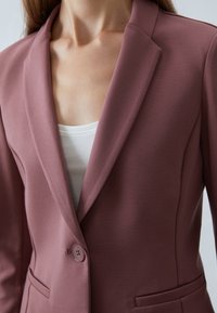 Rosa blazer med spetsig krage, enkel knappstängning och sidofickor. Slät textur och skräddarsydd passform. Buren över en vit topp.