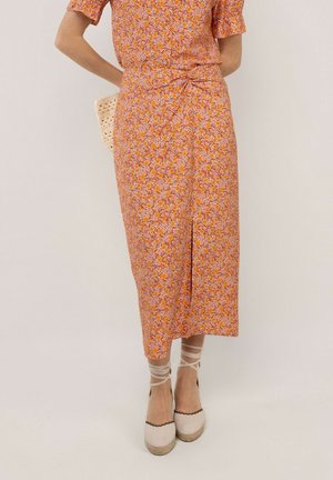 Mujer con vestido midi rosa con estampado floral y abertura frontal, zapatos tipo esparteña crema con ataduras en el tobillo, y sostiene un bolso tejido detrás de la espalda.