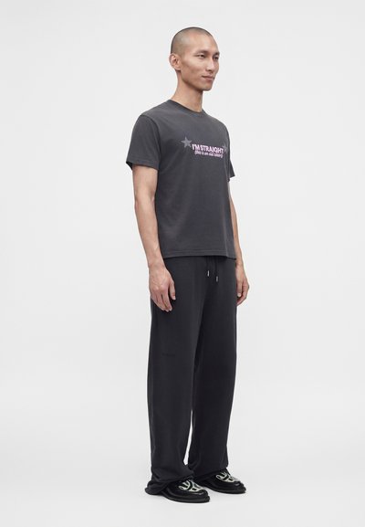 Άνδρας όρθιος, ντυμένος με μαύρο T-shirt "I'M STRAIGHT (this is an old shirt)", φαρδιά μαύρη παντελόνια και μαύρα παπούτσια με λευκά σχέδια.