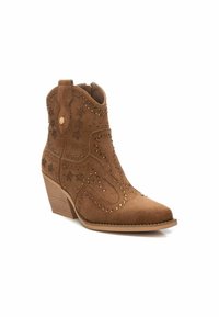 Bota de ante marrón hasta el tobillo con punta afilada, acentos de tachuelas decorativas y un tacón apilado. Presenta costuras de estilo western y tiradores laterales.