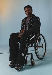 Czarny dres sportowy z białymi akcentami, szara koszulka i czarne buty sportowe. Osoba siedząca na wózku inwalidzkim na jasnoniebieskim tle.