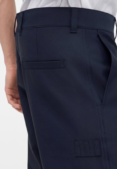 Gros plan sur un pantalon bleu marine foncé avec passants pour ceinture, poches latérales et arrière, porté avec une chemise gris clair rentrée.