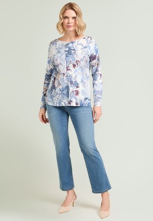 Kobieta stojąca w bluzce z długim rękawem w kwiaty, jasnoniebieskich jeansach o prostych nogawkach i beżowych butach na wysokim obcasie na jednolitym tle.