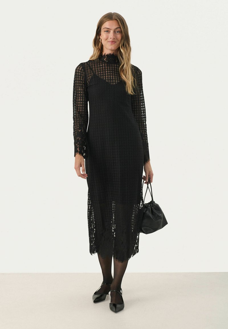 Schwarzes Kleid, strukturiertes Gewebe, lange Spitzenärmel und gewellter Saum. Kombiniert mit einer kleinen schwarzen Tasche und spitzen Ballerinas.