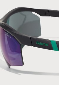 Polaroid-aurinkolasit mustalla kehyställä, violetit linssit ja vihreä korostus. Ominaisuuksiin kuuluu kulmikas muotoilu ja ULTRAPOLAR-merkintä sivussa.