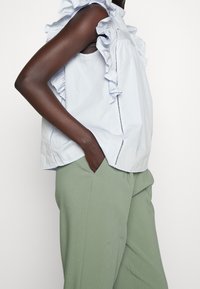 Lichtblauw mouwloos shirt met ruche schouderdetails en zijstiksels, gecombineerd met losse, lichtgroene broeken.