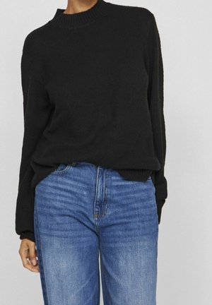 Personne portant un pull noir à manches longues rentré dans un jean bleu taille haute, debout devant un fond clair uni.