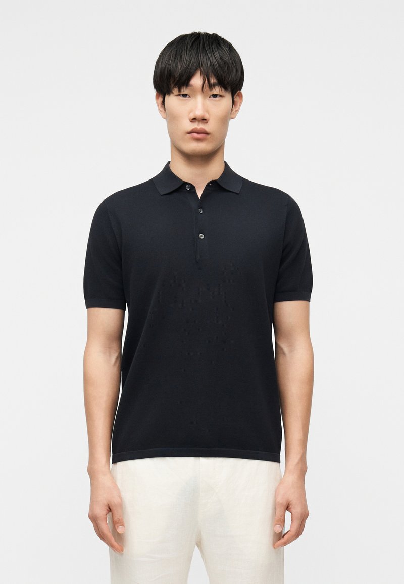 Homme portant un polo noir à manches courtes et un pantalon blanc, debout devant un fond gris uni.