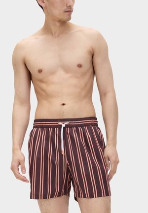 Homme portant un short de bain marron avec des rayures verticales roses et un cordon blanc, debout devant un fond uni.
