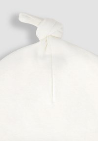 Valge kudumine müts, millel on ümar ülaosa ja seotud sõlm kroonil, esitleb pehmet tekstuuri ja minimalistlikku disaini.