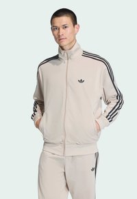 Chaqueta de chándal beige con cremallera, con rayas laterales negras y logo. Presenta un cuello alto y puños acanalados, combinada con pantalones a juego.