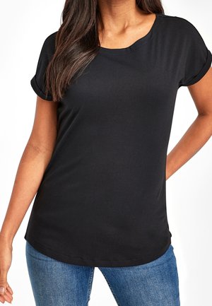 Kobieta ubrana w jednolity czarny t-shirt z krótkim rękawem z mankietami i niebieskie dżinsy, stojąca z jedną ręką za plecami.