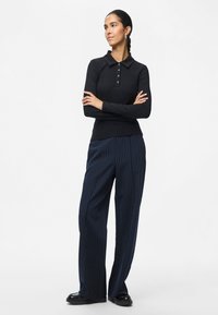 Camisa de polo de manga larga negra de canalé con detalles de botones, combinada con pantalones de pierna ancha a rayas verticales en azul marino y zapatos negros pulidos.