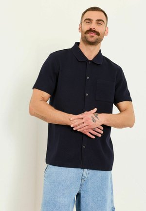 Mann mit Schnurrbart und Tattoos trägt ein schwarzes kurzärmeliges Hemd mit Knöpfen und helle blaue Jeans, steht vor einem einfarbigen weißen Hintergrund.