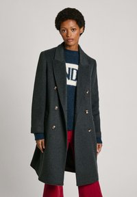 Manteau en laine double-breasted gris foncé avec deux poches avant, col cranté et boutons dorés. Porté par-dessus un pull en tricot bleu et un pantalon rouge.