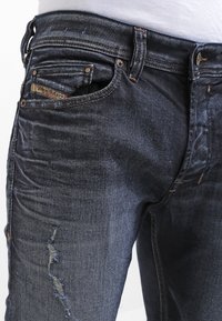 Mörkblå denimjeans med en sliten design som har synliga whiskers och fransade lappar, standard femficksdesign och metallaccenter.