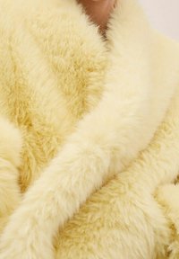 Osoba nosząca miękki, puszysty jasnobeżowy płaszcz z futra ekologicznego, pokazująca teksturę i szczegóły kołnierza.