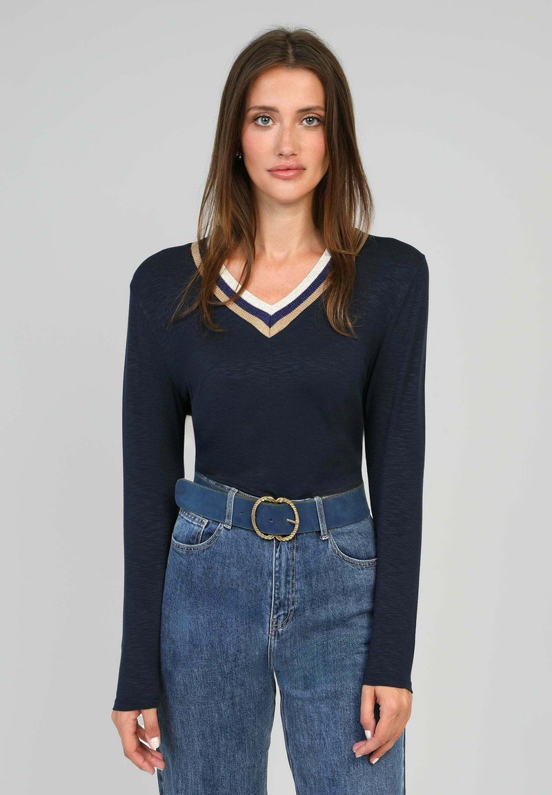 Haut à manches longues marine avec un col en V, orné d'accents rayés crème, marine et dorés. Associé à un jean en denim bleu taille haute et une ceinture bleue.