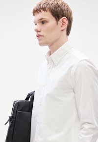 Λευκό πουκάμισο με γιακά και κουμπιά, με ένα μικρό λογότυπο. Το άτομο κρατάει μια μαύρη δερμάτινη τσάντα με φερμουάρ και χερούλι.