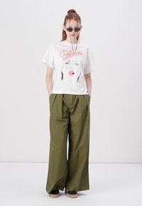 White graphic t-shirt featuring a face and "Blondie" text, paired with high-waisted olive green wide-leg pants, and black shoes.