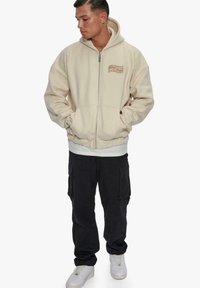 Beige zip-up hoodie with a kangaroo pocket, ribbed cuffs, and a graphic detail. Paired with black cargo pants and white sneakers.