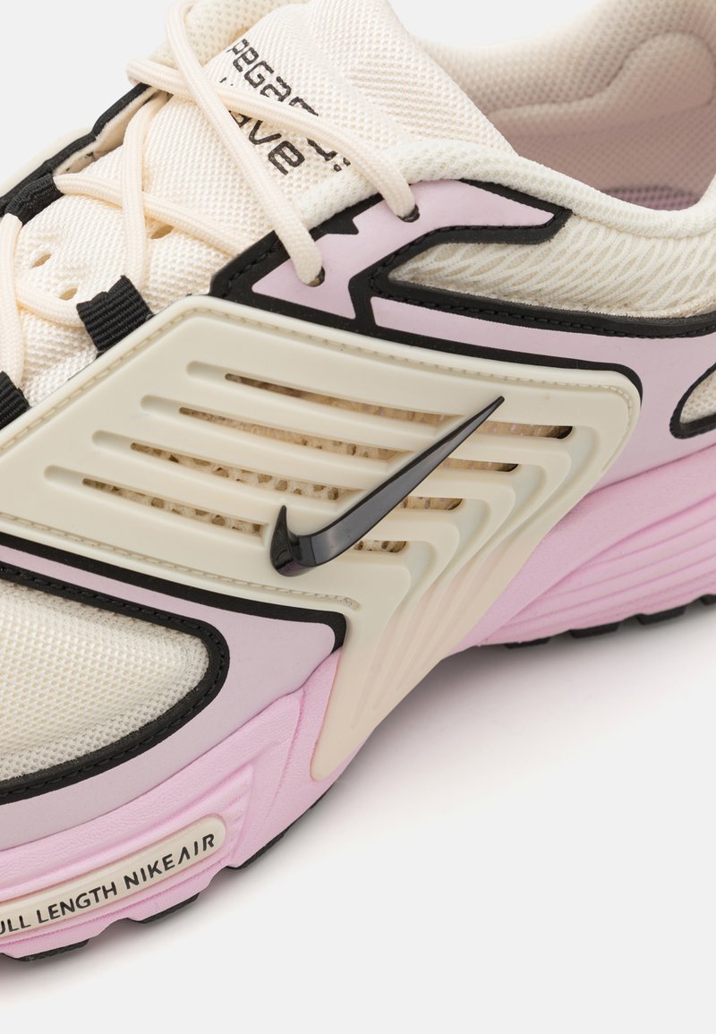 Zapatilla de estilo con parte superior de malla crema, acentos en rosa, detalles en negro y un prominente logo de swoosh negro en el costado.