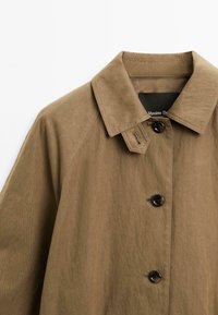 Brun letvægts trenchcoat med klassisk krave, knaplukning og tekstiler. Har en enkelt knapdetalje på kraven.