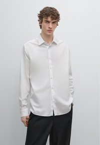 Witte blouse met lange mouwen en knopen, gemaakt van lichtgewicht stof. Voorzien van een klassieke kraag en een rechte snit, gedragen met een donkere broek.