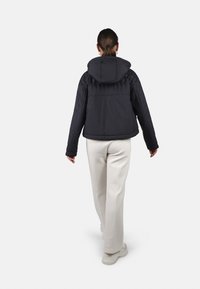 Giubbotto imbottito nero con cappuccio, caratterizzato da una finitura liscia e polsini a costine. Indossato con pantaloni chiari e sneakers.