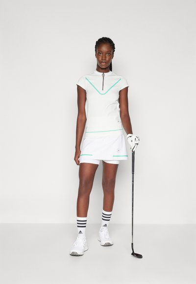 Fehér golfruházat, amely rövid ujjú, türkiz részletekkel díszített felsőből áll cipzáras gallérral, hozzá illő szoknyával és sportcipővel.