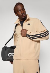 Veste zippée beige avec des rayures noires sur les manches ; matériau doux. Sac de sport noir avec un logo blanc tenu dans une main.