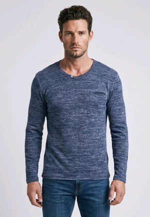 Hombre con el pelo corto y rizado que lleva una camisa azul ajustada de manga larga con textura y vaqueros azules, de pie frente a un fondo claro y liso.