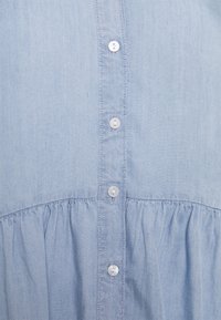 Camicia azzurro chiaro con bottoni, realizzata in un tessuto morbido, con orlo arricciato e sei bottoni bianchi lungo la apertura. Texture liscia.