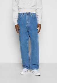 Jeans de denim azul claro con corte recto, costuras visibles, cinco bolsillos y una cinturilla con botón y cremallera. Combinados con zapatillas blancas.