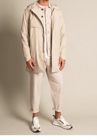 Ljus beige vattentät jacka med huva, dragkedja och framficka, bärs över en ljus skjorta med knappar och beige byxor. Vita sneakers.