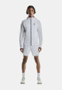 Veste de sport blanche avec une fermeture éclair noire, assortie à un short blanc. Tissu lisse, design minimaliste, avec un logo sur la poitrine.